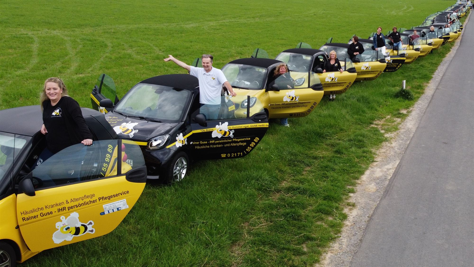 Drohnenaufnahme: lange Reihe gelb-schwarzer Elektro-Dienstwagen mit Bienen-Logo auf einer Wiese, Mitarbeitende winken
